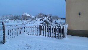 Un balcón cubierto de nieve con valla de madera, una casa al fondo y un cielo despejado.