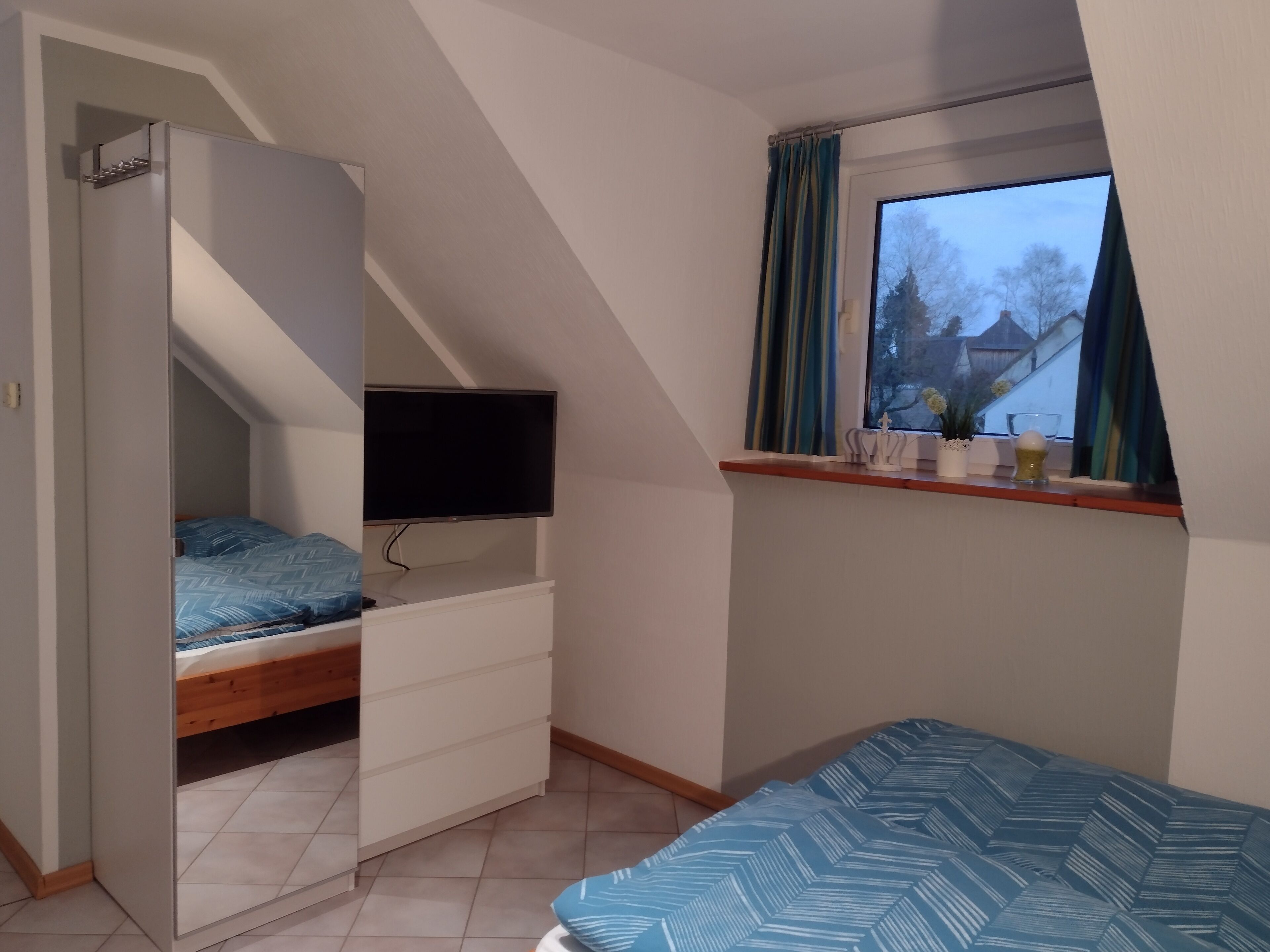Une chambre à coucher avec un lit, un téléviseur fixé au mur, une fenêtre avec des rideaux et une petite étagère sur laquelle sont posés quelques objets.
