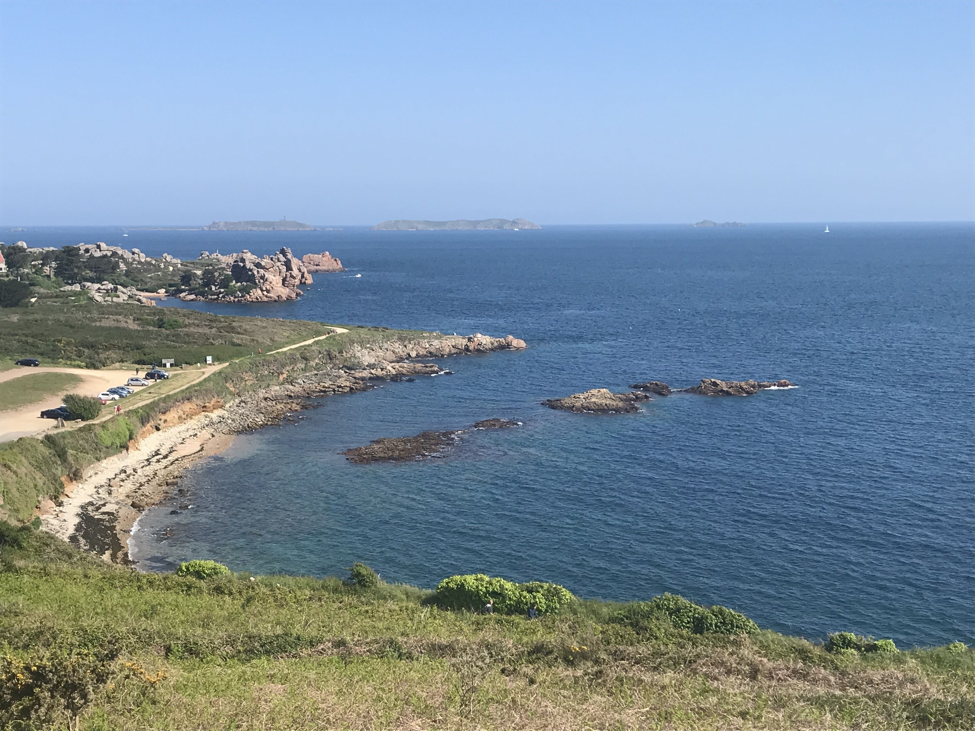 A coastal landscape with rocky outcrops, a clear blue sea, and a path leading to a parking area.