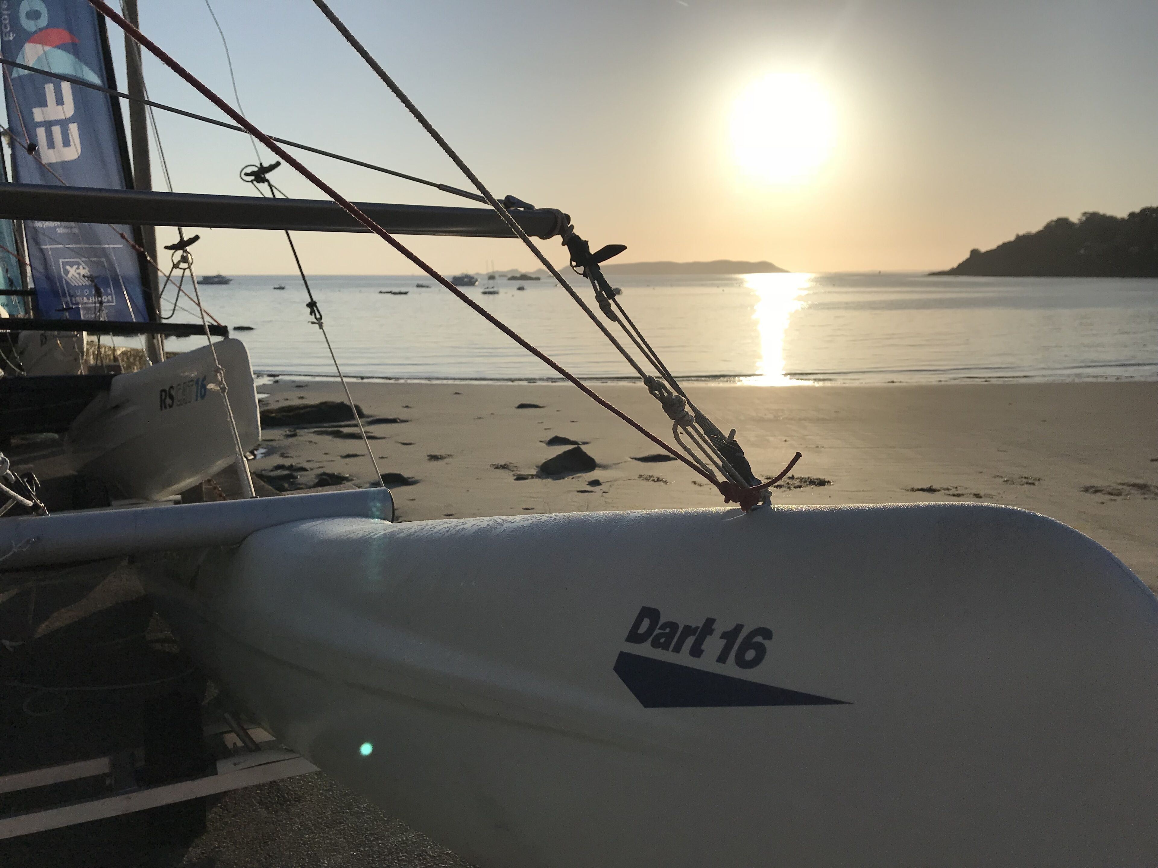 A sailboat with the name "Dart 16" on its hull, docked at a beach with the sun setting in the background.