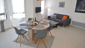 A modern living room with a round wooden table, grey chairs, a grey sofa with orange pillows, a floor lamp, and a flat-screen TV mounted on the wall. - Apartment *** approved 2018 - Trestraou Beach (Perros-Guirec)