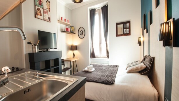 A compact hotel room with a kitchenette, a bed with a blanket, a TV, a small table with a lamp, and a window with curtains.