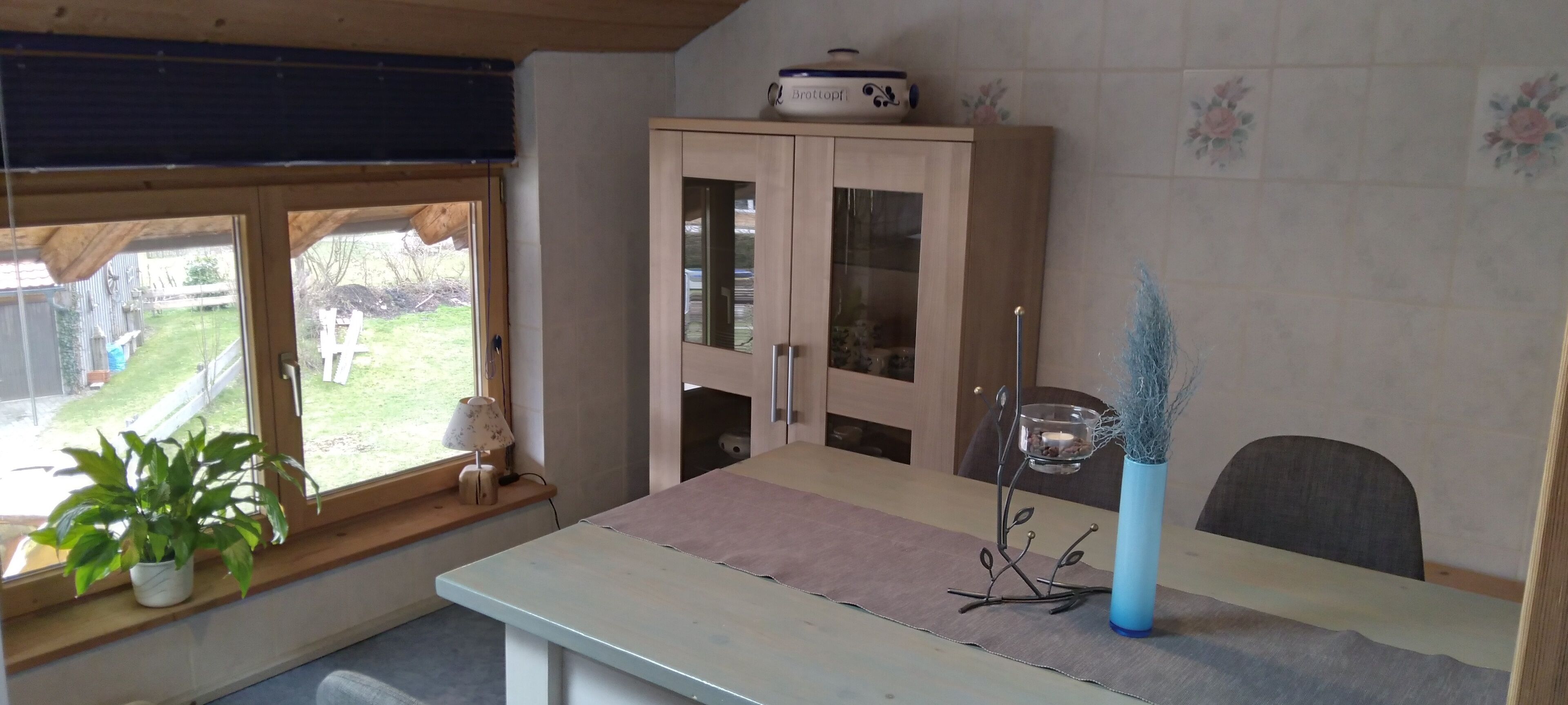 Ein Zimmer mit großem Fenster, einem Holzschrank, einem Tisch mit Stoffauflage und einer Zimmerpflanze in einem Topf sowie einer Pflanze in einer Vase.