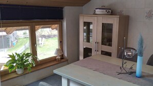 Een kamer met een groot raam, een houten kast, een tafel met een tafelkleed, een potplant en een vaas met planten.