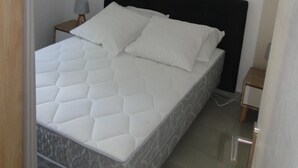 Uma cama bem arrumada com colchão e travesseiros brancos, uma mesinha de cabeceira de madeira com abajur e uma pequena mesa de madeira ao lado da cama.