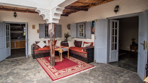 A cozy room with a wooden ceiling, a red patterned rug, a sofa with cushions, and a small table. - Villa Tigmin Eden with traditional Moroccan decor. (Essaouira)