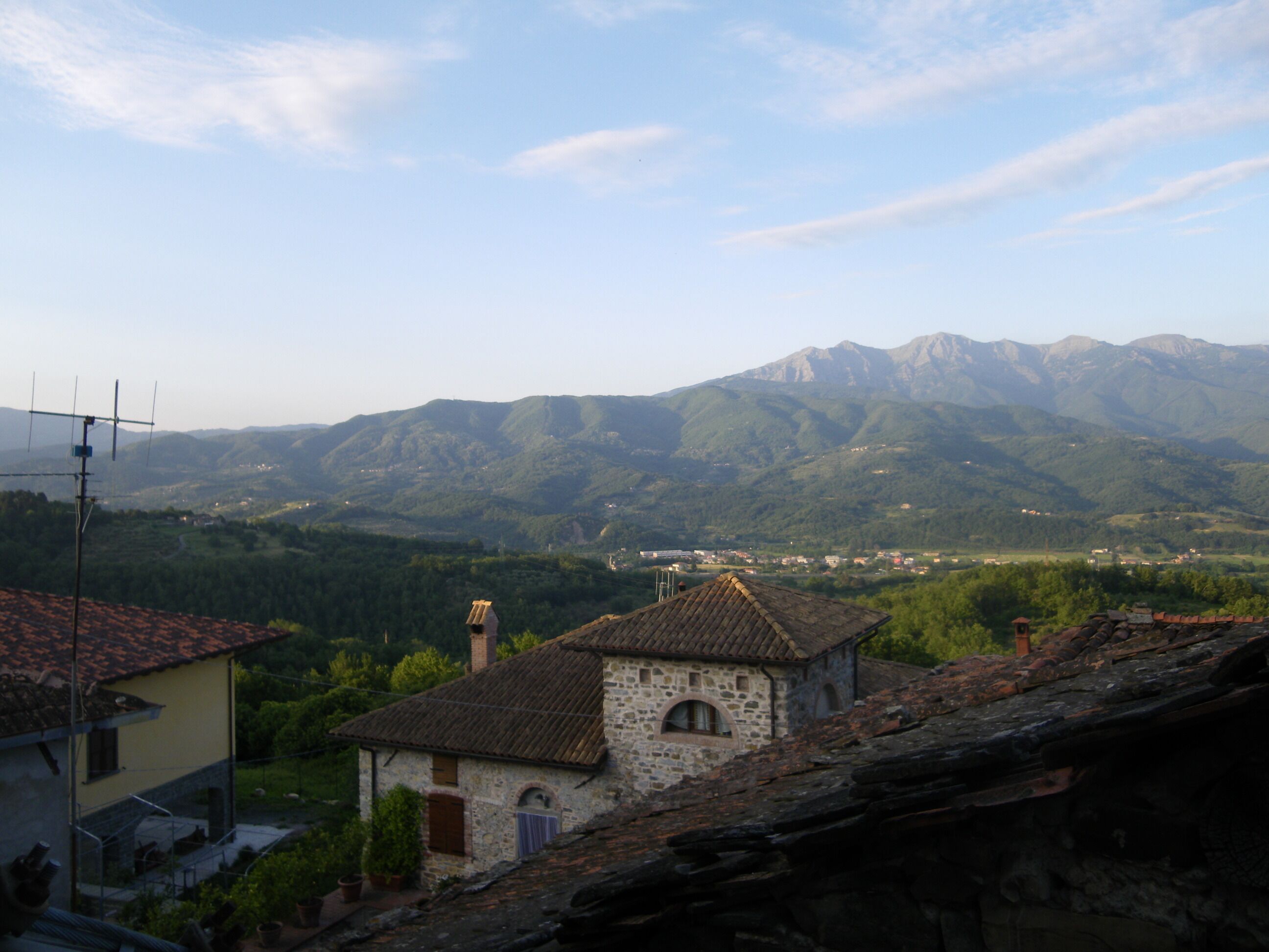A stone building with a tiled roof, surrounded by lush greenery and mountains in the background.