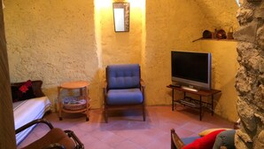 Een gezellige kamer met een stenen muur, een blauwe fauteuil, een houten bijzettafeltje met een televisie en een klein rond tafeltje met een mand.
