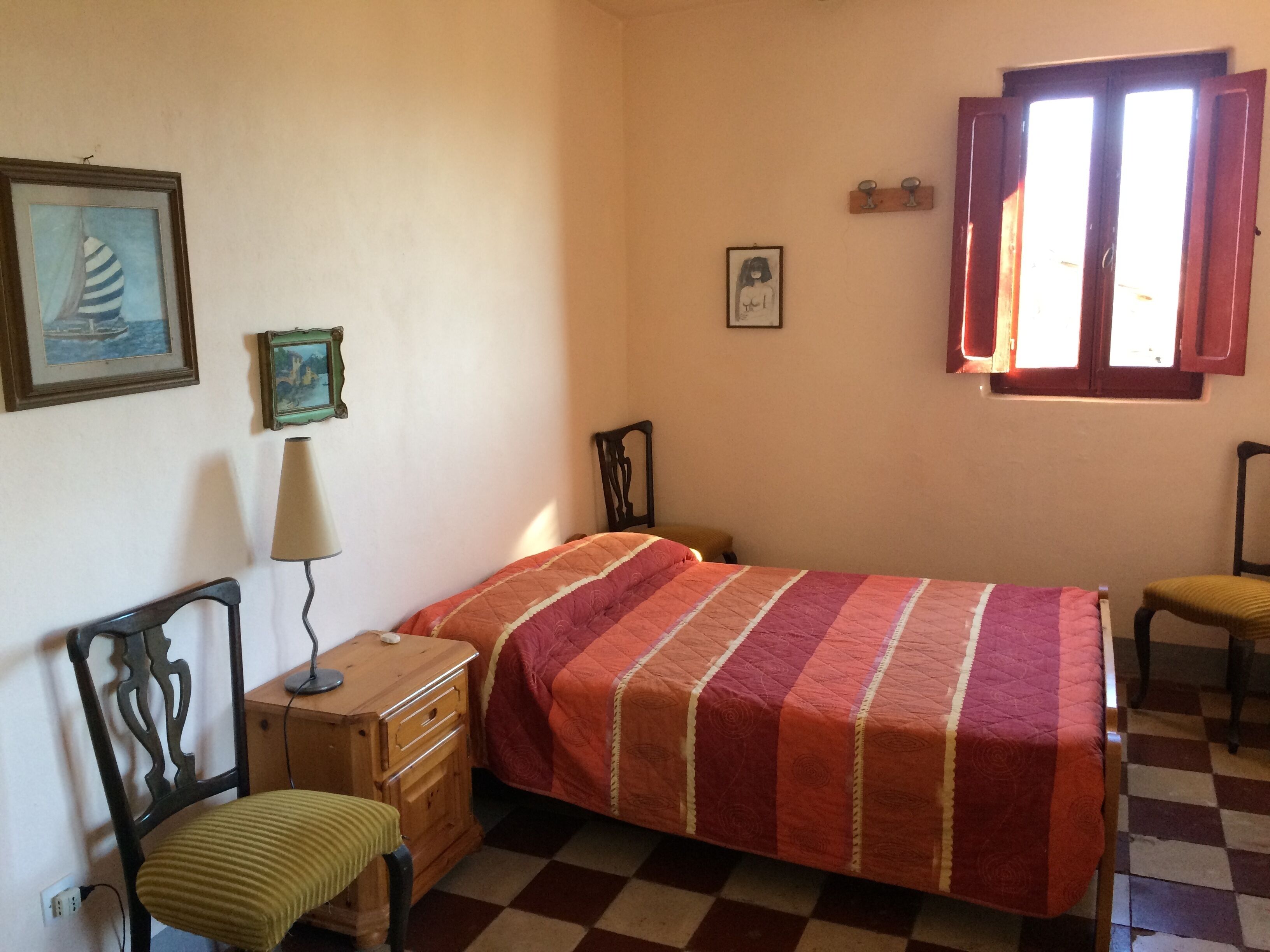 A bedroom with a checkered floor, a bed with a striped bedspread, a wooden nightstand, a chair, a lamp, and two framed pictures on the wall.