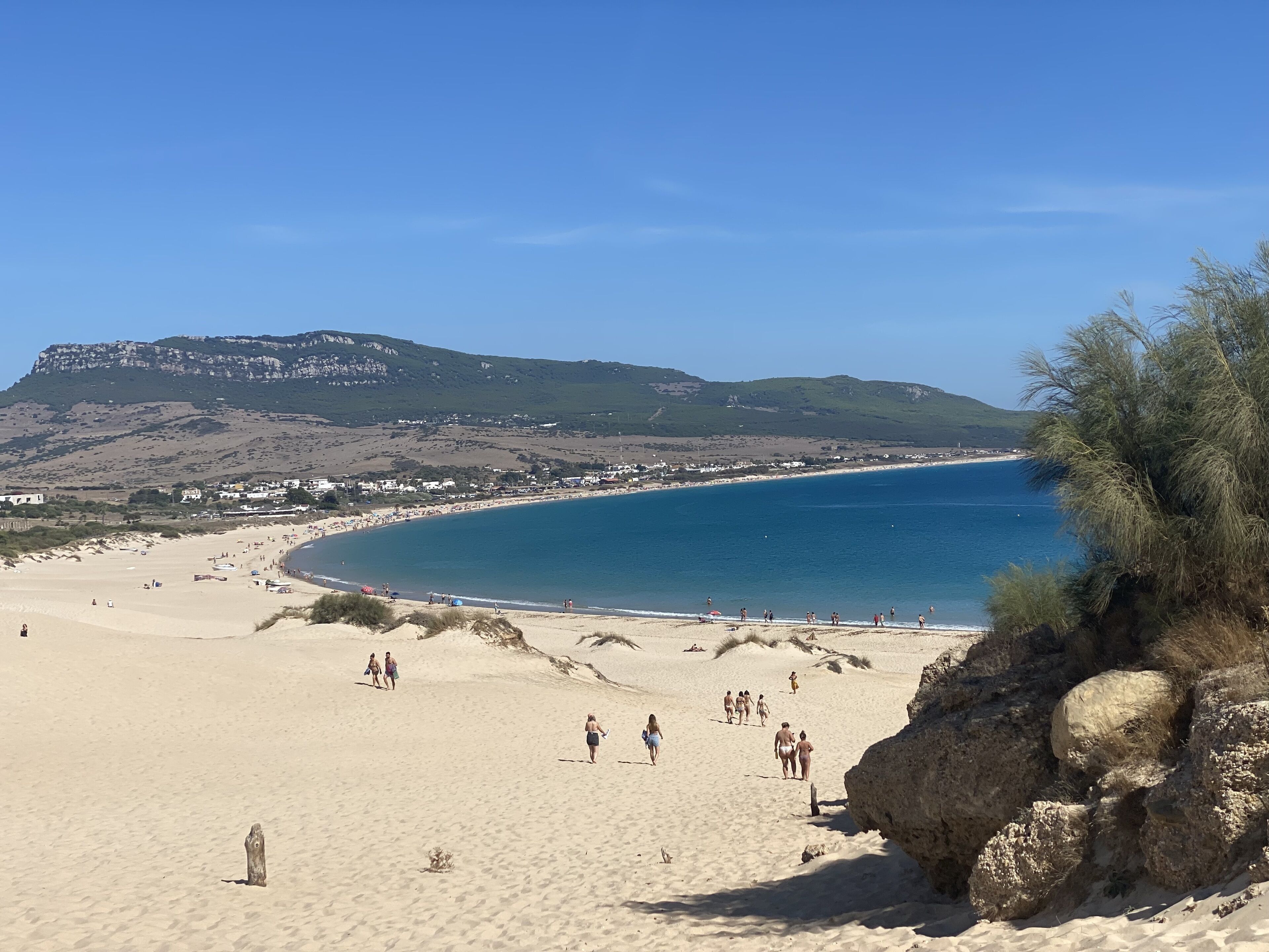 A beach with a clear view of the sea, a rocky outcrop, and a distant hill with buildings.