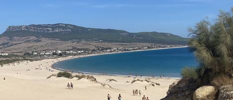 A beach with a clear view of the sea, a rocky outcrop, and a distant hill with buildings.