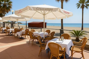Outdoor tables with white tablecloths and wicker chairs, under large umbrellas with 'Cruzcampo' branding, overlooking a beach.