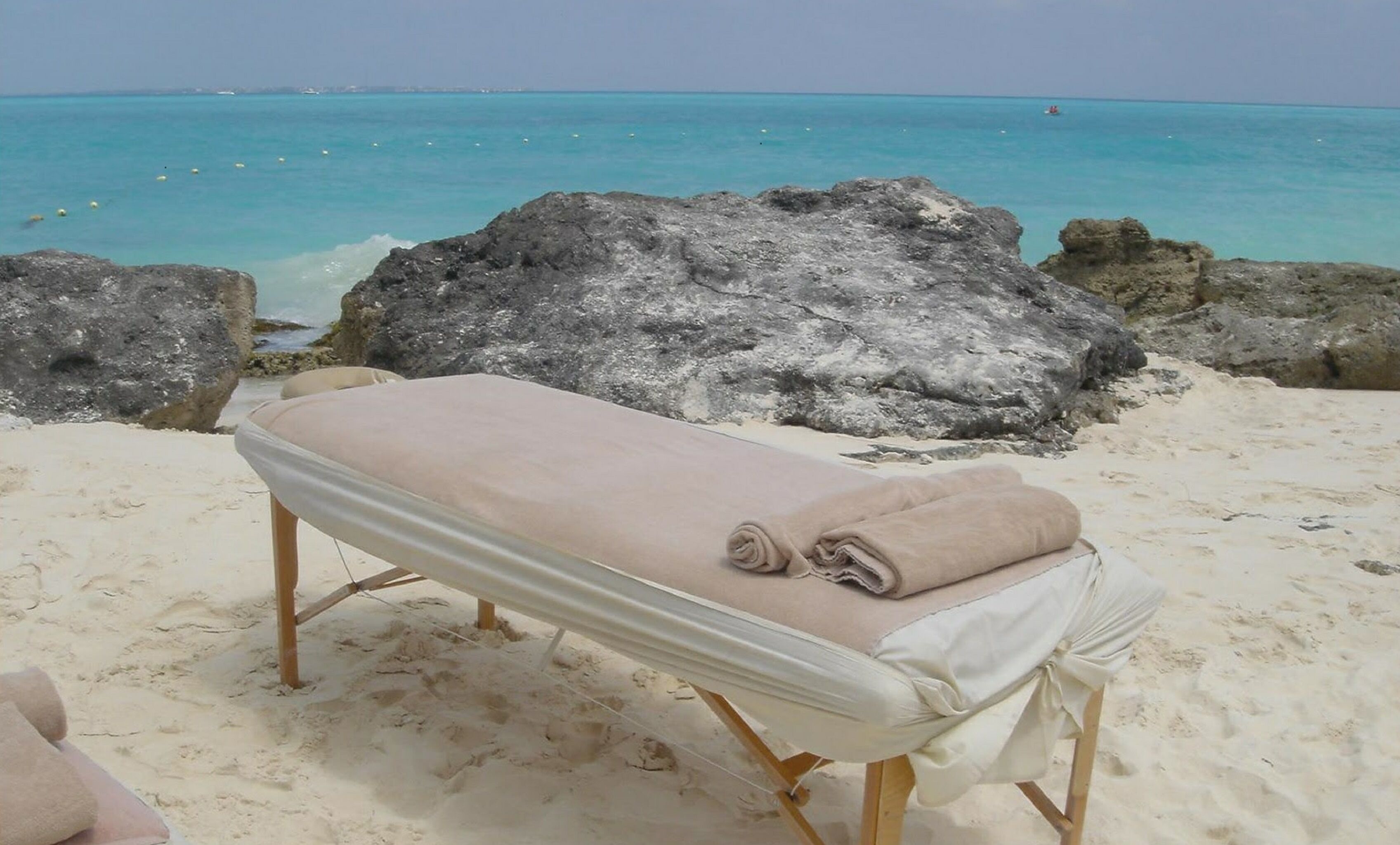 Una sedia a sdraio in riva al mare, con un asciugamano color beige, sullo sfondo di una costa rocciosa e un mare color turchese.