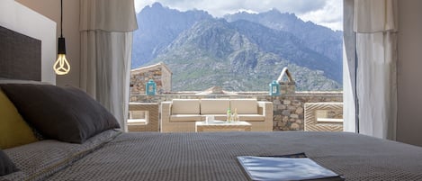 Quarto com cama, mesa de cabeceira e vista para montanhas pela janela.