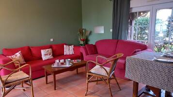 A cozy living room with a red sofa, wicker chairs, a wooden coffee table, and a dining table with a patterned tablecloth.