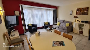 A living room with a TV, sofa, chairs, a dining table, and a kitchen area.