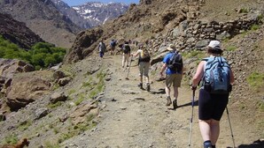 Um grupo de caminhantes sobe por uma trilha rochosa na montanha, sob um céu claro e com picos nevados ao longe.
