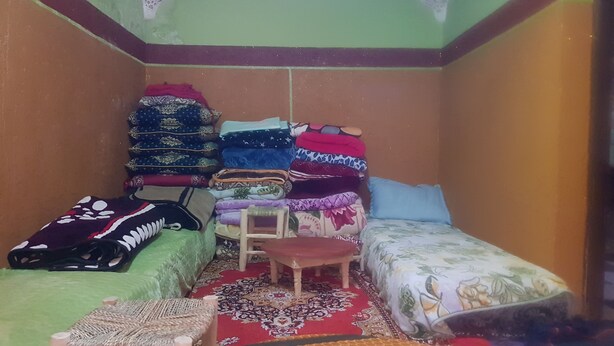 Quarto pequeno e acolhedor com duas camas de solteiro, um banquinho de madeira e uma cadeira.