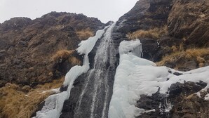 Uma cascata despencando sobre um penhasco rochoso, cercada por grama seca.