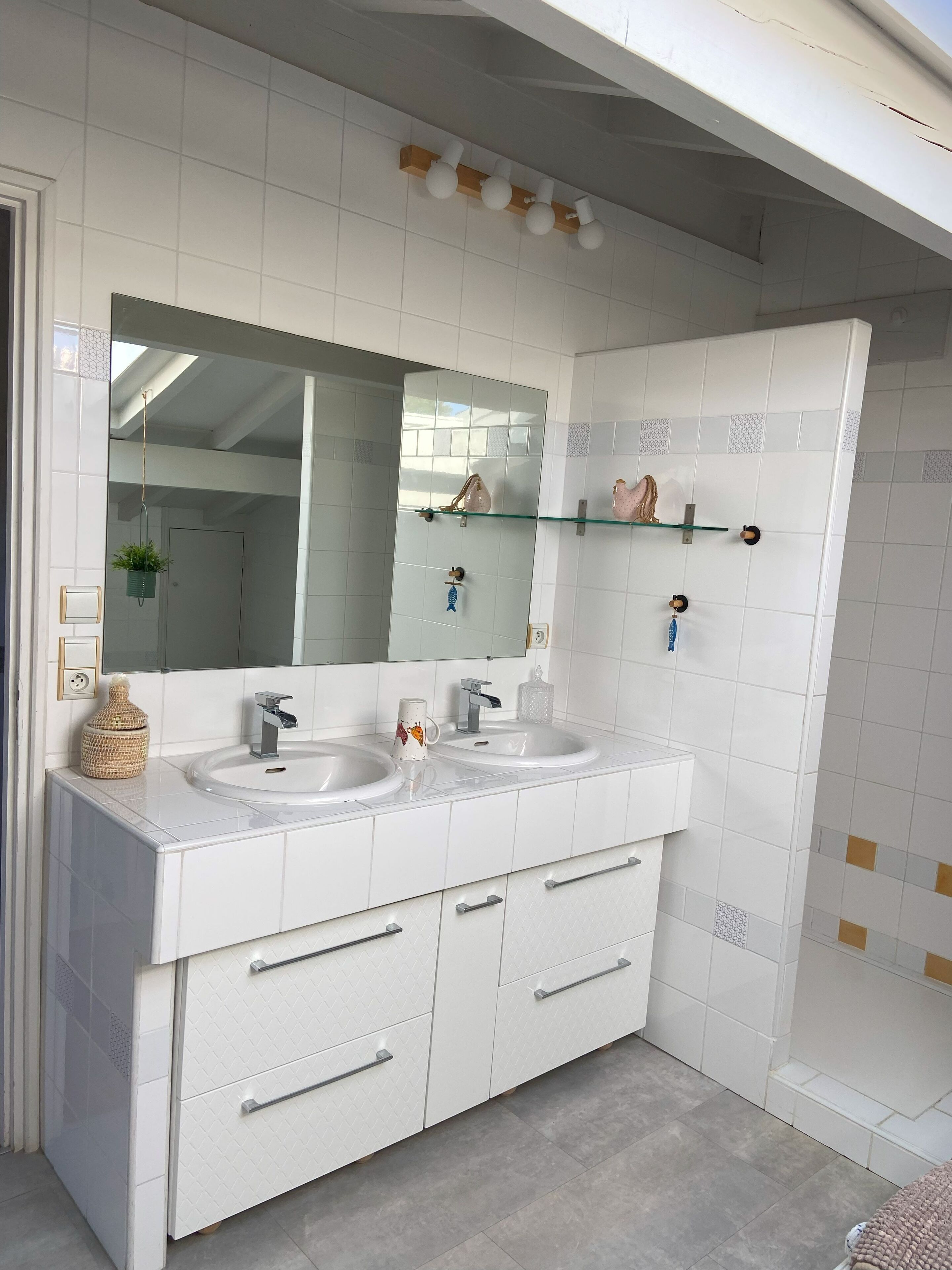 Een badkamer met een wastafelmeubel met dubbele wastafel, een grote spiegel en een glazen plank met decoratieve objecten.