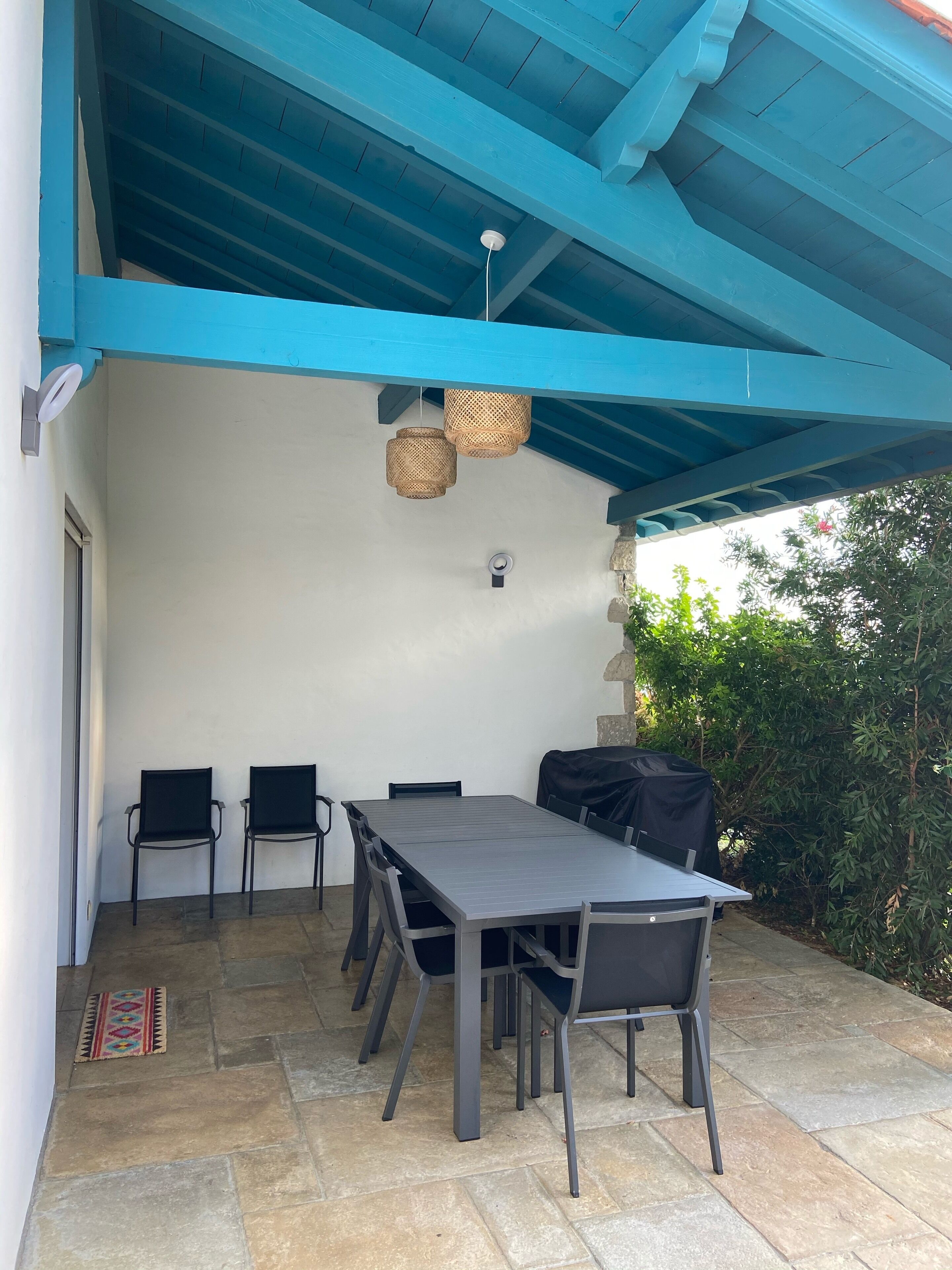 Une terrasse extérieure avec un plafond bleu, une table avec des chaises, un barbecue et un luminaire suspendu.