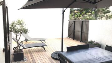 Une terrasse en bois avec une table et des chaises, un transat et un grand parasol.