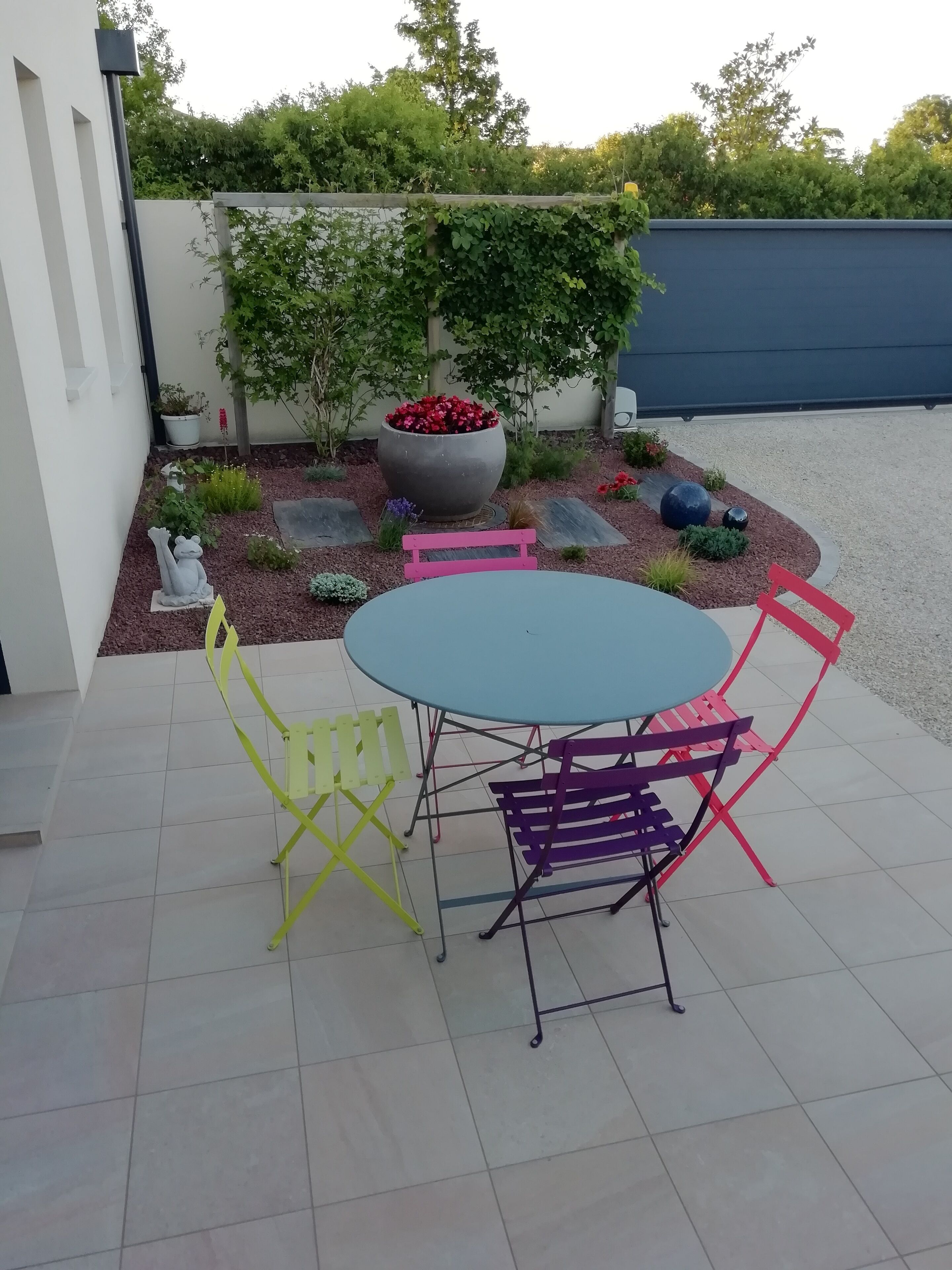A patio with a round table and colorful chairs, surrounded by a garden with a large planter and decorative rocks.