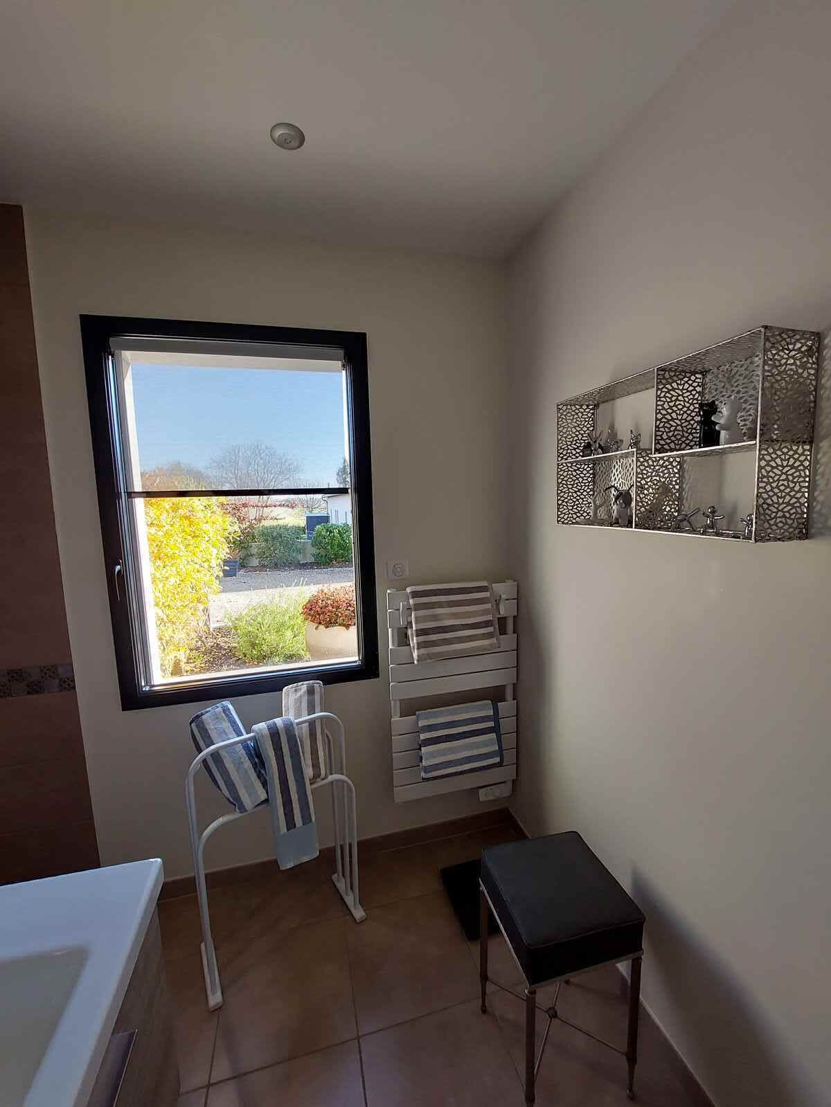 A bathroom with a large window, a towel rack, a chair, a small table, and a decorative mirror.
