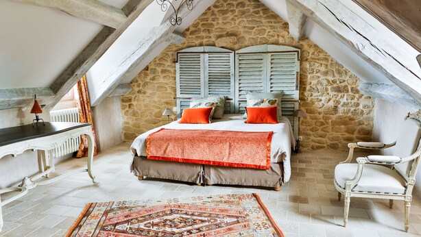 Een slaapkamer met een stenen muur, een bed met oranje kussens, een witte stoel en een vloerkleed met een patroon.