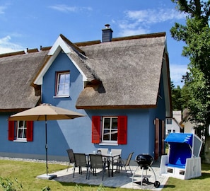 A house with a thatched roof, a patio with a table and chairs, and a barbecue grill.