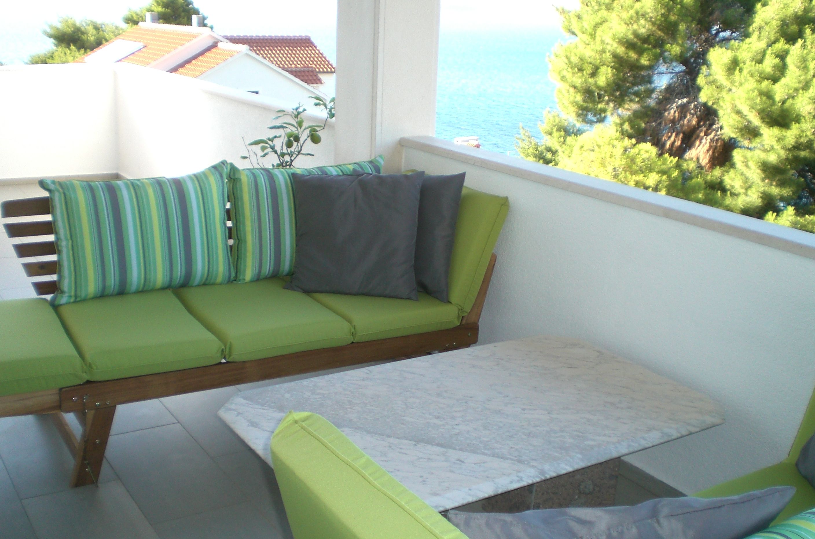 A balcony with a green sofa, a small table, and a view of the sea and trees.