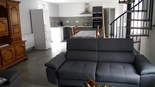 A modern living room with a grey sofa, a wooden cabinet, and a glass coffee table. In the background, there is a kitchen with a built-in oven and a staircase leading to another level. - renovated holiday home - sleeps 8 (FLAVIAC)