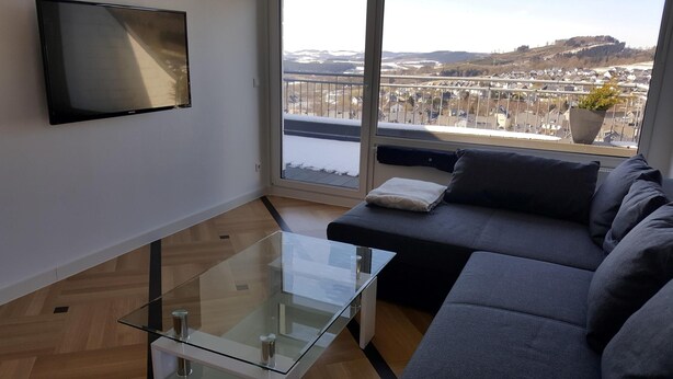 Un salon moderne avec une table basse en verre, un canapé gris et un téléviseur fixé au mur.