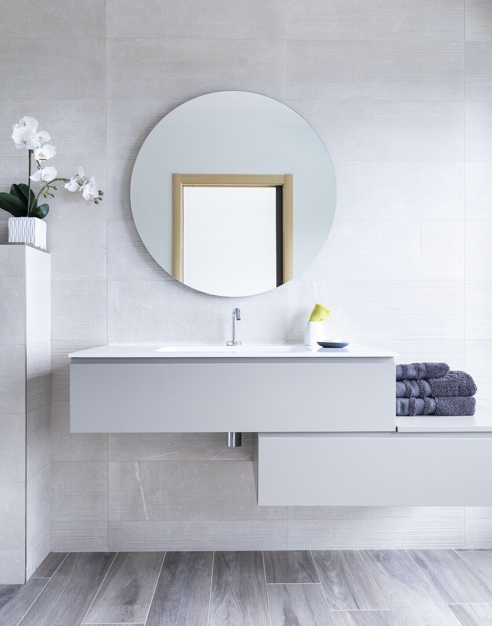Ein modernes Badezimmer mit einem weißen Waschtisch, einem runden Spiegel und einer kleinen Orchidee im Topf.