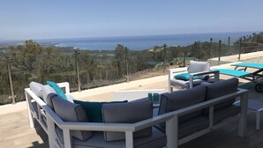 A terrace with a white sofa and armchairs, a small table, and a view of the sea and hills.
