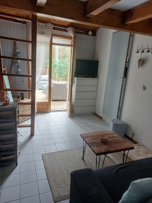 A compact living space with a tiled floor, a wooden table, a sofa, a television, and a door leading to an outdoor area with a balcony.