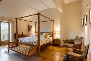 Una cama con dosel de madera, un banco, dos mesitas de noche con lámparas y sillones de mimbre.