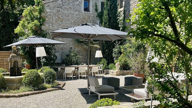 Een terras buiten met stenen tafels en stoelen, parasols en een fontein.