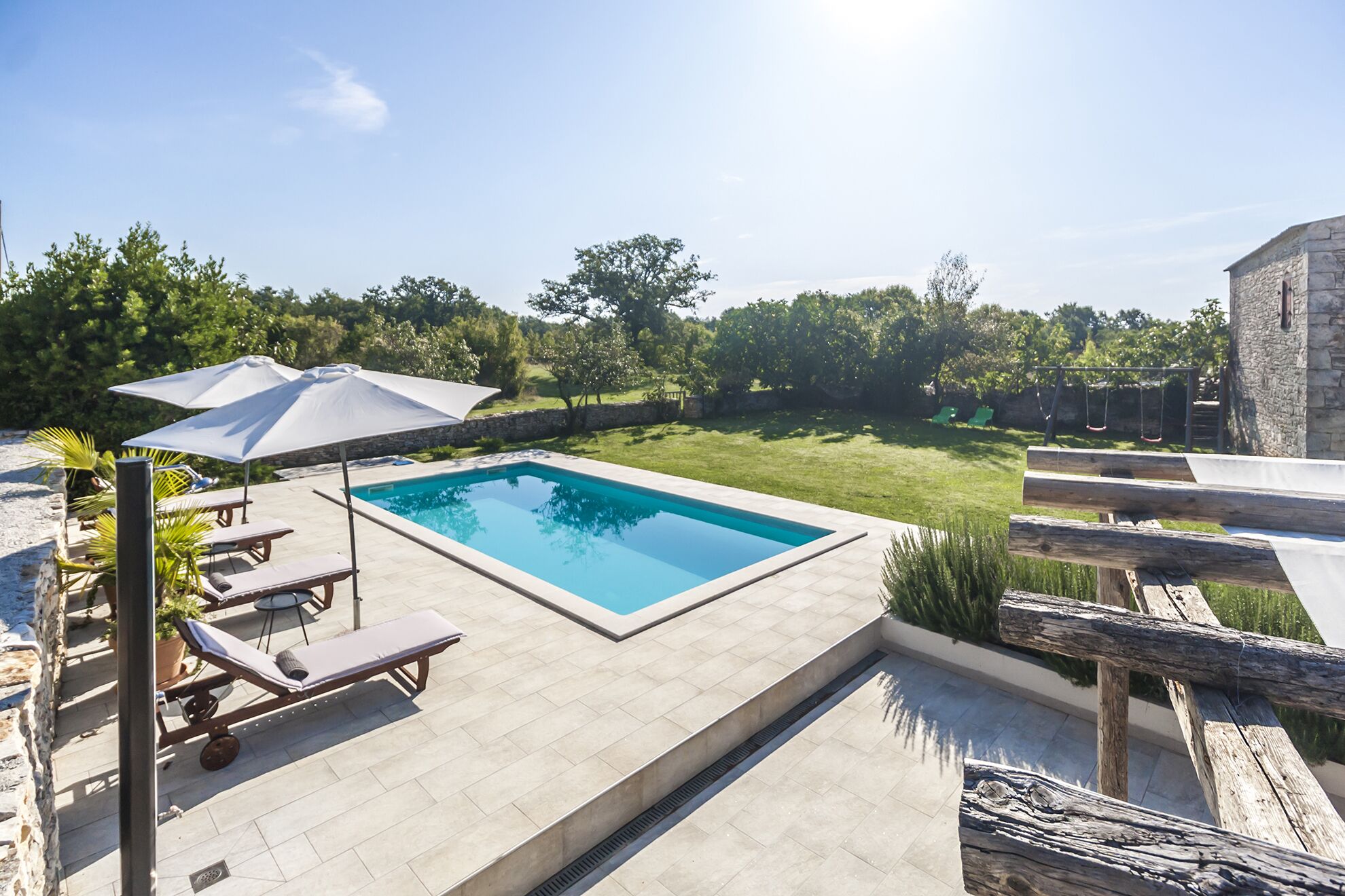 Piscina retangular com um deck revestido de azulejos, rodeada por um muro de pedra e um deck de madeira.