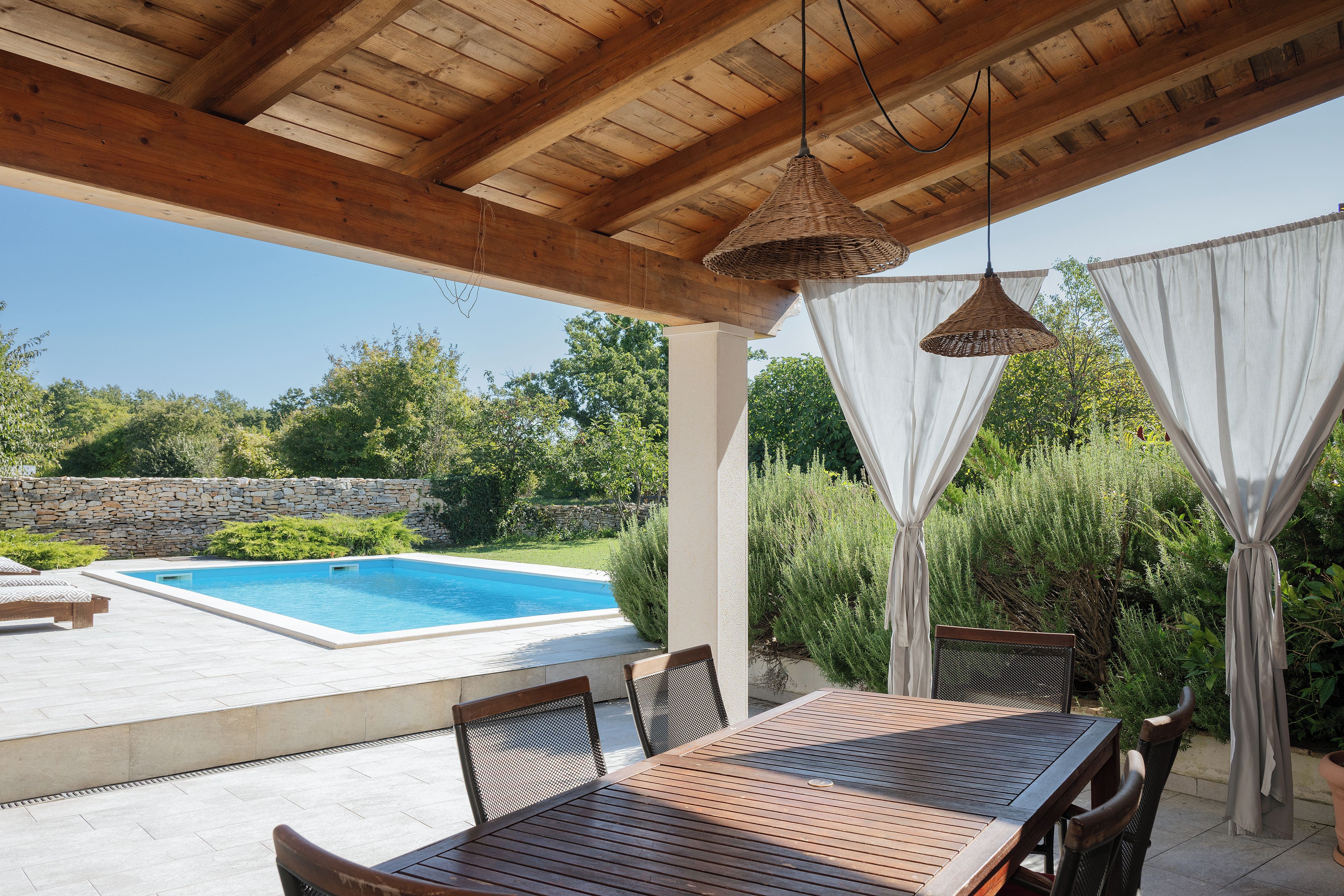 A covered outdoor area with a pool, wooden furniture, and a stone wall.