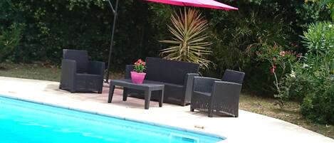 Zona de piscina amb seients, cadires, una taula, un parasol rosa i una planta en test.