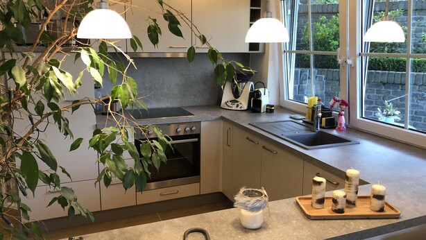 Een moderne keuken met een ingebouwde kookplaat, oven en wastafel. Er zijn hangplanten, een houten dienblad met kaarsen en een groot raam met uitzicht op het omliggende landschap.