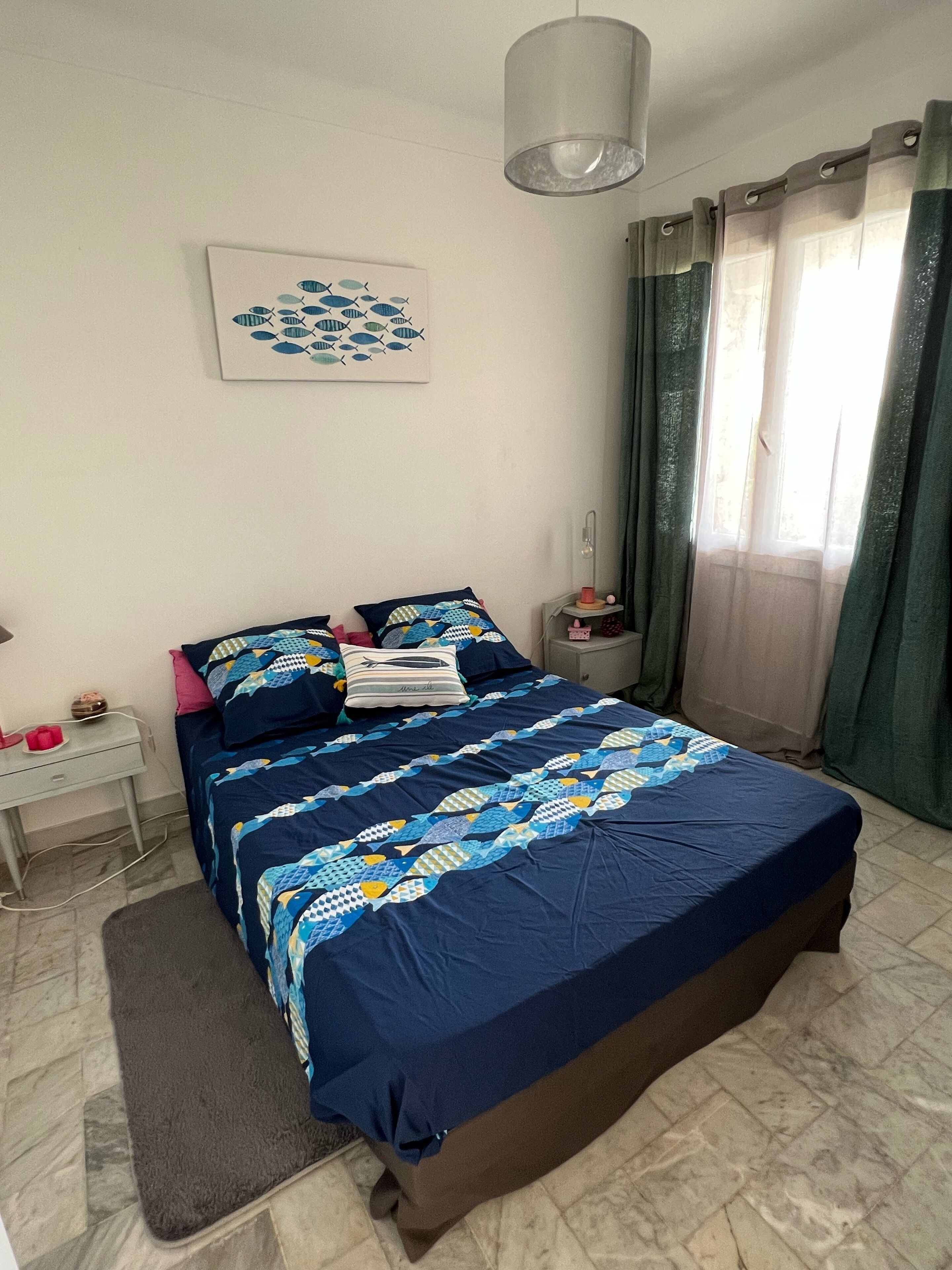 A bedroom with a large bed, a bedside table, a lamp, a window with curtains, and a wall-mounted artwork.