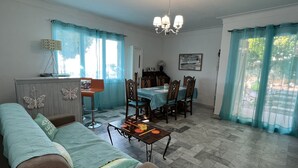 A living room with a sofa, a dining table with chairs, a chandelier, and a kitchen area with a refrigerator. - 3 room apartment on the ground floor of a villa with sea view (Mandelieu-La-Napoule)