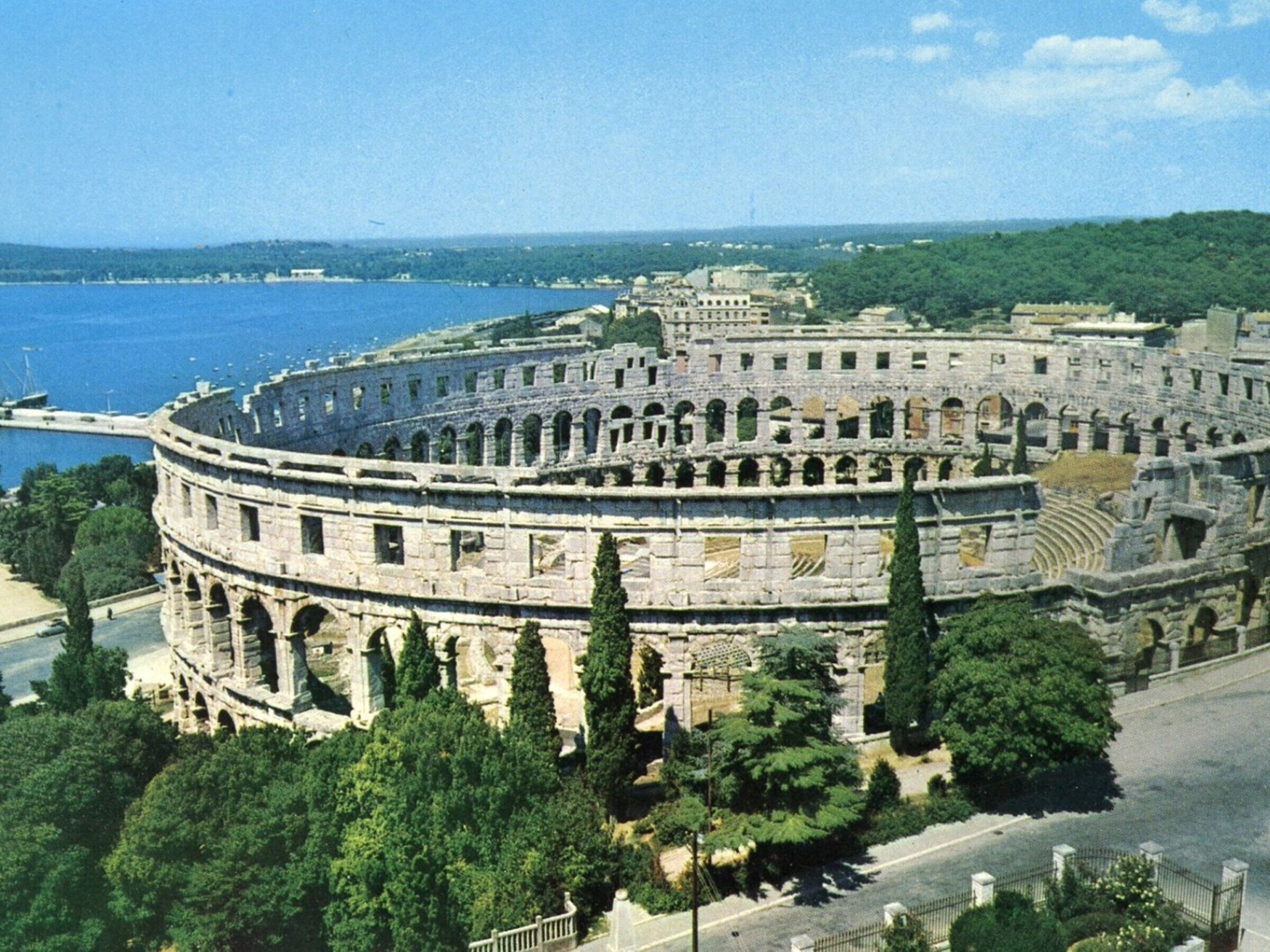Ein antikes Amphitheater mit mehreren Sitzstufen, umgeben von Bäumen und einem Gewässer.