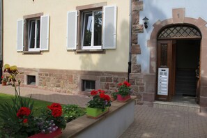 Ein Gebäude mit Steinwänden, einer Holztür mit der Aufschrift '1' darüber und Kübelpflanzen mit roten Blüten vor der Fassade.
