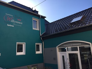 Un bâtiment de couleur turquoise avec un toit en tuiles, une porte portant l'inscription « Bodenbelage Tapeten Farben » et une fenêtre sur laquelle on peut lire « www.westerburger-farbenwelt.de ».