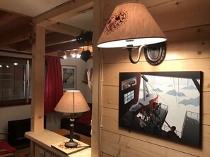 Una habitación con pared de madera, una mesa con lámpara, un cuadro enmarcado y un ventanal con cortina roja.