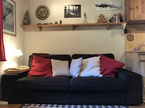 A dark-colored sofa with four pillows in a living room. A wooden shelf above the sofa displays various items including a clock, a small Christmas tree, and a picture. - Appartement 65m2 esprit Chalet. Parking. Balcon (Cauterets)