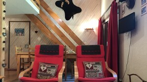 Ein gemütliches Zimmer mit schrägverdeckter Decke, zwei rote Polsterstühle mit gemusterten Kissen, ein Holztisch und ein Fernseher an der Wand.